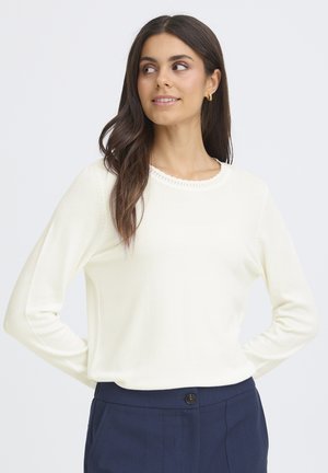 Cremfarbener Strickpullover mit Rundhalsausschnitt und dekorativen Nähten. Lange Ärmel, weiche Textur, kombiniert mit dunkelblauen Hosen.