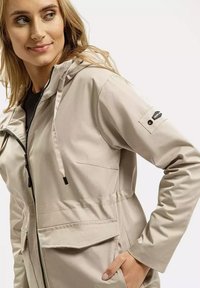 Jasno beżowa wodoodporna kurtka z kapturem, z zamkiem z przodu, elastycznymi mankietami oraz dużą przednią kieszenią z klapką. Czarne akcenty z logo.