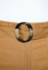 Béžový oděv s páskem z tan látky a černobílou oválnou sponou, která je umístěna ve středu.