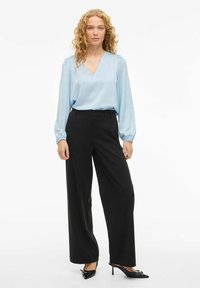 Blouse en soie bleu clair avec un col en V et des manches bouffantes, associée à un pantalon noir à jambes larges et des escarpins noirs ornés d'accents décoratifs.