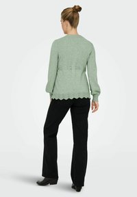 Zeleni pleteni pulover z dolgimi rokavi, rebrastim robom in pentljami na hrbtni strani. Skupaj z črnimi širokimi hlačami in čevlji s peto.