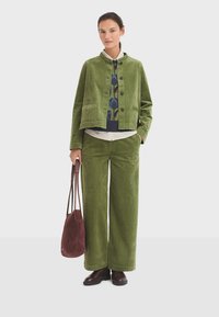 Green corduroy jacket with round collar and two front pockets, paired with matching wide trousers, worn with a patterned sweater and a burgundy bag.