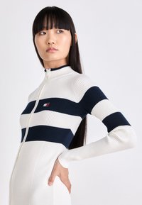 Body a manica lunga con zip, aderente e rigato in crema con strisce orizzontali blu scuro; presenta un colletto alto e una piccola toppa con logo sul petto.