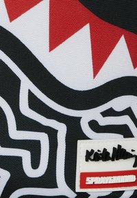 Tejido negro con patrones abstractos en blanco y rojo, con una etiqueta blanca que muestra "Keith Haring" y "SPRAYGROUND" en texto negro y rojo.