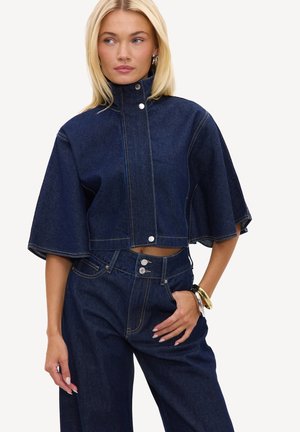 Femme blonde portant une veste cape en denim bleu foncé avec des boutons-pression et un jean large taille haute assorti, accessoirisée avec des bracelets.