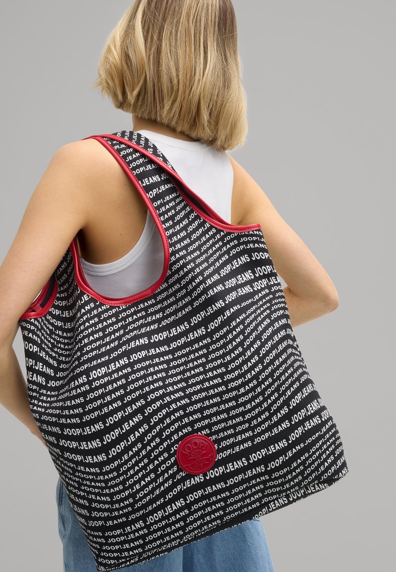 Czarna torba na ramię z białym, powtarzającym się napisem "JOOP!JEANS", czerwonymi akcentami i okrągłą łatką z logo. Zaprojektowana z szerokimi, wygodnymi paskami.
