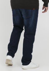 Jeans de mezclilla azul oscuro exhibidos desde la parte trasera, con un corte relajado y dos bolsillos traseros. Combinados con zapatillas blancas para crear un contraste.