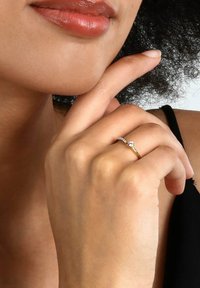 Guld Ring med en liten central klar sten och tvinnad banddetalj. Hand som vilar nära hakan, vilket framhäver ringens design och glittrande effekt.