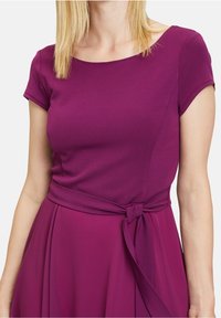 Fialové šaty s přiléhavým korzetem a krátkými rukávy, se stahováním v pase a splývavou sukní. Hladká tkanina s mírným leskem.