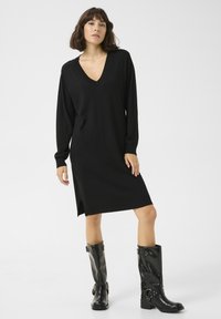 Schwarzes langärmliges Kleid mit V-Ausschnitt, seitlichen Schlitzen und lockerer Passform, aus einem weichen Stoff gefertigt. Kombiniert mit schwarzen kniehohen Stiefeln.