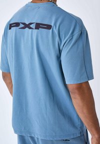 Camiseta de algodón azul claro con mangas cortas, cuello redondo y un gran logotipo negro "PXP" en la parte posterior. Tela texturizada, corte relajado.