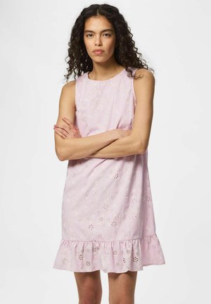 Kvinde med krøllet hår iført ærmeløs lyserød kjole med blomsterhulmønster og flæsekant, stående med armene over kors.