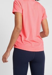 Korálově pruhované sportovní tričko s kulatým výstřihem a krátkými rukávy, spárované s námořnickými legínami. Tkanina se zdá být hladká a lehká.