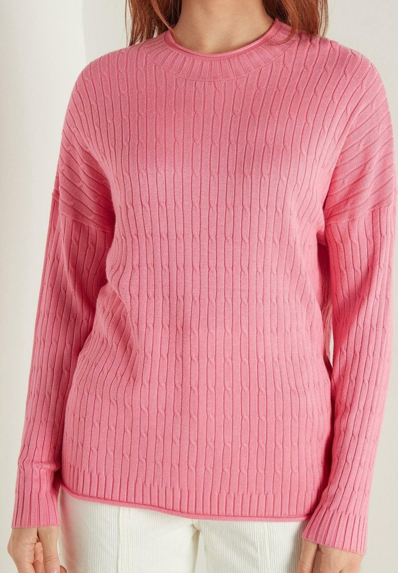 Femme portant un pull rose torsadé à manches longues avec poignets et ourlet côtelés, associé à un pantalon blanc.
