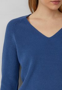 Niebieski sweter z dzianiny o dekolcie w kształcie litery V, z prążkowaną teksturą i długimi rękawami, mocno przycięty, aby uwydatnić szczegóły tkaniny.