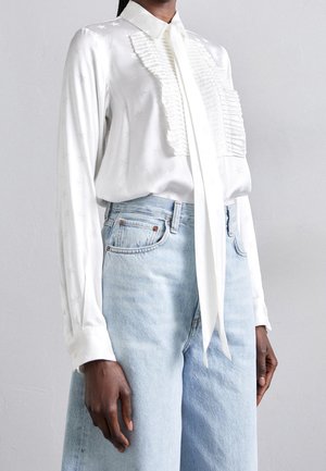 Witte satijnen blouse met een gestructureerde ruchesdetail aan de voorkant, lange mouwen en een nektie. Gecombineerd met lichtblauwe denim short.