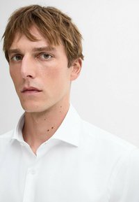 Weißes, knopfbares Hemd aus Baumwolle mit spitzem Kragen und glatter Textur. Das Gesicht des Models ist teilweise sichtbar, vor neutralem Hintergrund.