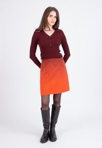 Femme debout, les mains derrière le dos, portant un haut à manches longues bordeaux, une jupe orange, des collants noirs et des bottes noires jusqu'au genou.
