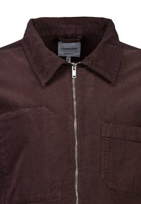 Chaqueta de pana marrón con cuello puntiagudo, cierre de cremallera plateado y dos bolsillos en el pecho. Tejido estructurado y texturizado con un diseño de ajuste moderno.