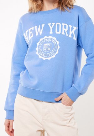 Donna che indossa una felpa azzurro chiaro con scritta bianca "New York" ed emblema a tema baseball, abbinata a pantaloni beige chiaro.