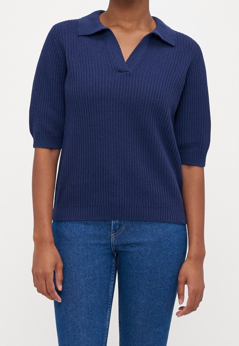 Personne portant un pull en maille bleu marine à manches courtes avec col, associé à un jean en denim bleu, debout devant un fond uni.