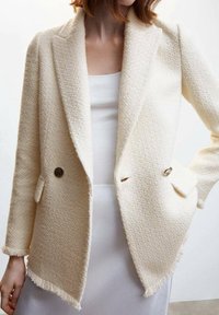 Cremfarbiger Blazer mit Reverskragen, doppelter Knopfleiste, sichtbaren Nähten an den Säumen und runden Metallknöpfen.