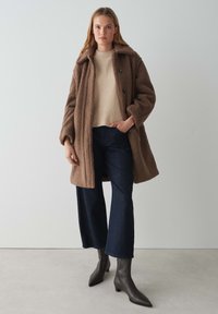 Manteau brun et duveteux avec un grand col et des boutons noirs, porté sur un pull beige clair et un jeans foncé évasé ; associé à des bottines grises.