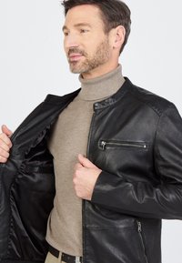 Man in een lichtbruine turtleneck die een open zwarte leren jas met ritszakken aanpast tegen een effen achtergrond.