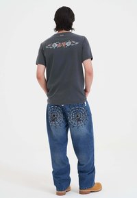 Szara koszulka z kwiatowym wzorem skrzydeł na plecach, zestawiona z niebieskimi dżinsami z haftem pajęczej sieci na kieszeniach. Brązowe obuwie.