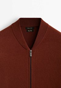 Bruine ritstrui met een geribde kraag, gemaakt van zacht materiaal. Beschikt over een zilveren rits en een label in de hals.