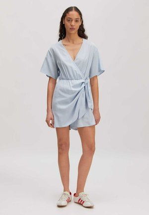 Frau mit lockigem Haar, die ein hellblaues Wickelkleid und weiße Sneakers mit roten Streifen trägt, steht vor einem schlichten Hintergrund.