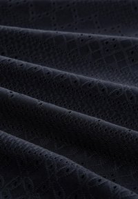 Tejido oscuro que presenta un patrón texturizado con pequeñas formas geométricas y agujeros espaciados uniformemente, creando un contraste sutil en la superficie.