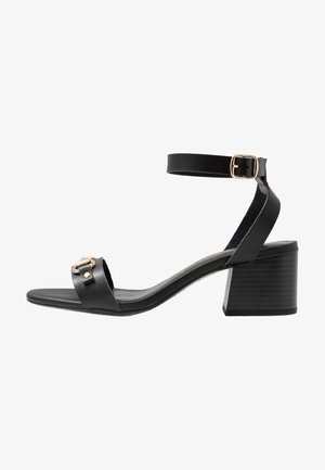 Schwarze Leder-High-Heel-Sandale mit Knöchelriemen, goldfarbenen Hardware-Akzenten und strukturiertem Blockabsatz. Offene Zehenpartie.