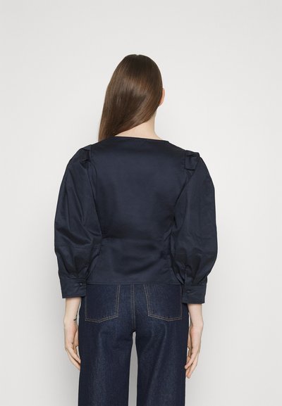 Ναυτικό μπλε σατέν τοπ με φουσκωμένα μανίκια, διαθέτοντας στρογγυλή λαιμόκοψη και εφαρμοστό σιλουέτα. Το ύφασμα φαίνεται λείο και γυαλιστερό.