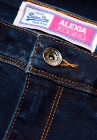 Tummansiniset farkkujeggingit, joissa on keltaset tikit, metallipainike ja etiketti, jossa lukee "SuperDry Alexia Jegging".