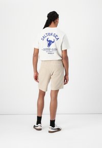 Outfit casual que incluye una camiseta crema con gráficos azules y pantalones cortos beiges. Los pantalones cortos tienen textura y un bolsillo trasero. Se completa con calcetines negros y zapatillas.