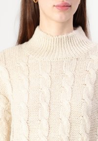 Pūkuotas kreminis turtleneck megztinis su kabeliniu raštu, corrugacija apykakle ir švelnia, jaukia medžiaga su švelniais storio svyravimais.
