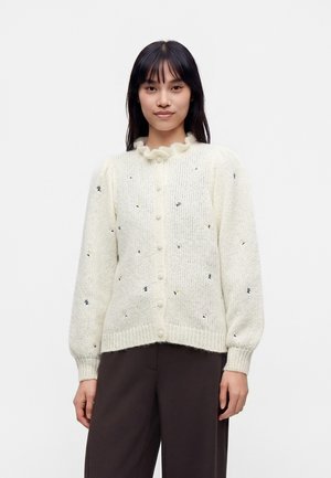 Ung kvinde bærer en cremefarvet strikket cardigan med flæset krave, knapper foran og små broderede detaljer, kombineret med mørkebrune bukser.
