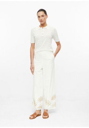 Femme portant un polo blanc à manches courtes et un pantalon blanc à jambes larges avec des motifs de feuilles dorées aux ourlets, associé à des sandales marron, debout.