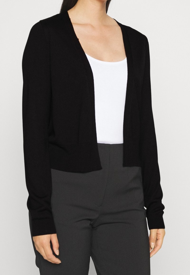 Cardigan nero corto in morbido materiale a maglia con maniche lunghe e scollatura a V. Abbinato a un top bianco e pantaloni grigi.