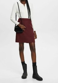 Haut à manches longues côtelé noir avec fermeture éclair, associé à une jupe bordeaux. Le modèle porte des bottines noires et tient un petit sac bandoulière noir.