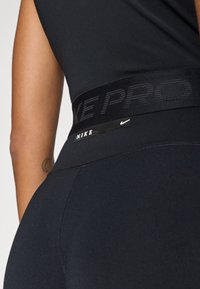 Leggings negre Nike Pro, cu o talie groasă și text ridicat "NIKE PRO" și un mic etichetă cu logo-ul Nike. Textură de țesătură netedă și ajustată.