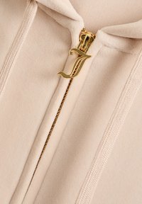 Lichtbeige ritsjack met een gouden ritslip met een ornate "J" ontwerp, zachte stofstructuur en ribbelaccenten langs de rits.