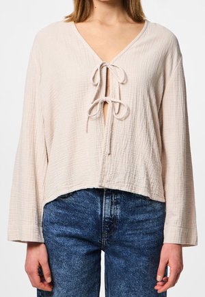 Kvinde iført en lysebeige, tekstureret bluse med lange ærmer og to bundne sløjfer foran, kombineret med blå denimjeans.