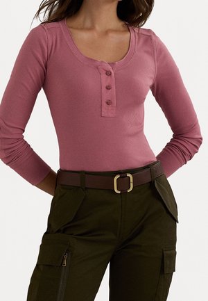 Woman wearing a dusty pink long-sleeve henley top tucked into dark olive cargo pants with a brown belt and gold buckle.