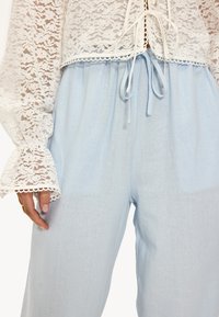 Blouse en dentelle avec des manches cloche longues, nouée à l'avant, associée à un pantalon large à cordon de serrage en bleu clair. Tissu lisse avec une texture subtile.