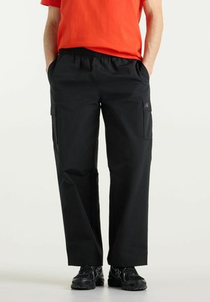 Persona che indossa pantaloni cargo neri con tasche laterali, scarpe da ginnastica nere e una maglietta arancione a maniche corte con le mani nelle tasche.