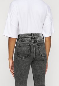 Jeans grises de cintura alta con una textura desgastada, cinco bolsillos y un parche de logo plateado en la cintura trasera. Camisa blanca holgada por encima.
