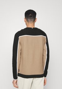 Maglione lavorato a maglia con corpo beige e maniche nere, caratterizzato da dettagli bianchi lungo la cucitura. Design con scollo rotondo, tessuto texturizzato.