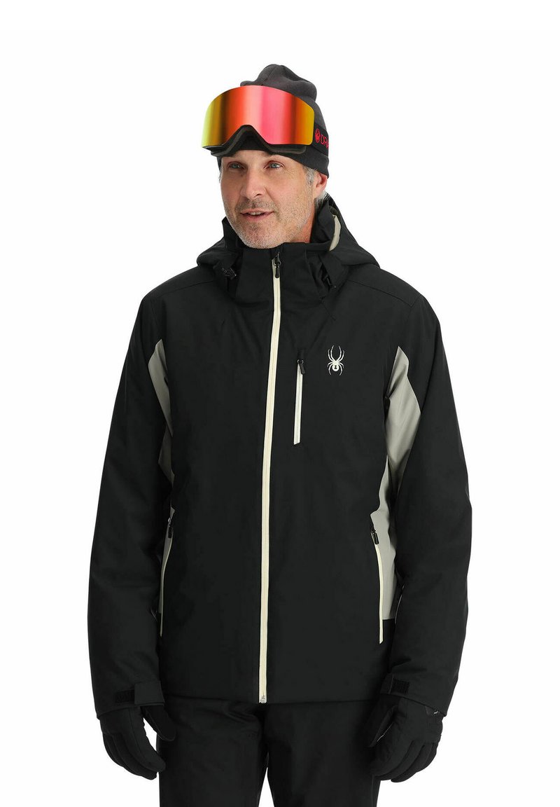 Schwarze isolierte Jacke mit grauen Akzenten, Frontreißverschluss und einem Spinnenlogo. Getragen mit einer Mütze und verspiegelten Skibrillen. Handschuhe und Skihosen sichtbar.